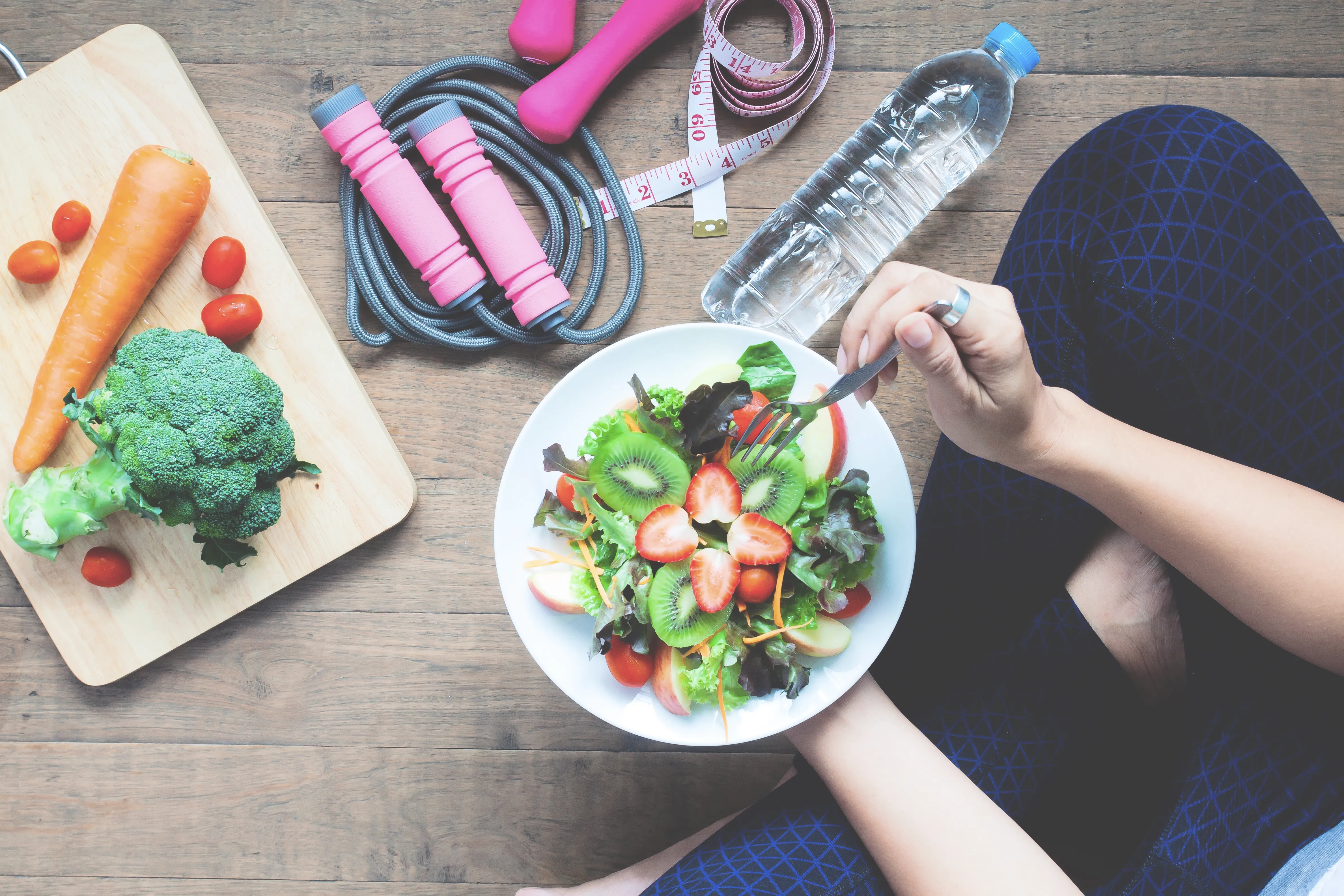 Healthy food, a skipping rope, water bottle and measuring tape arranged beside a person eating a salad
