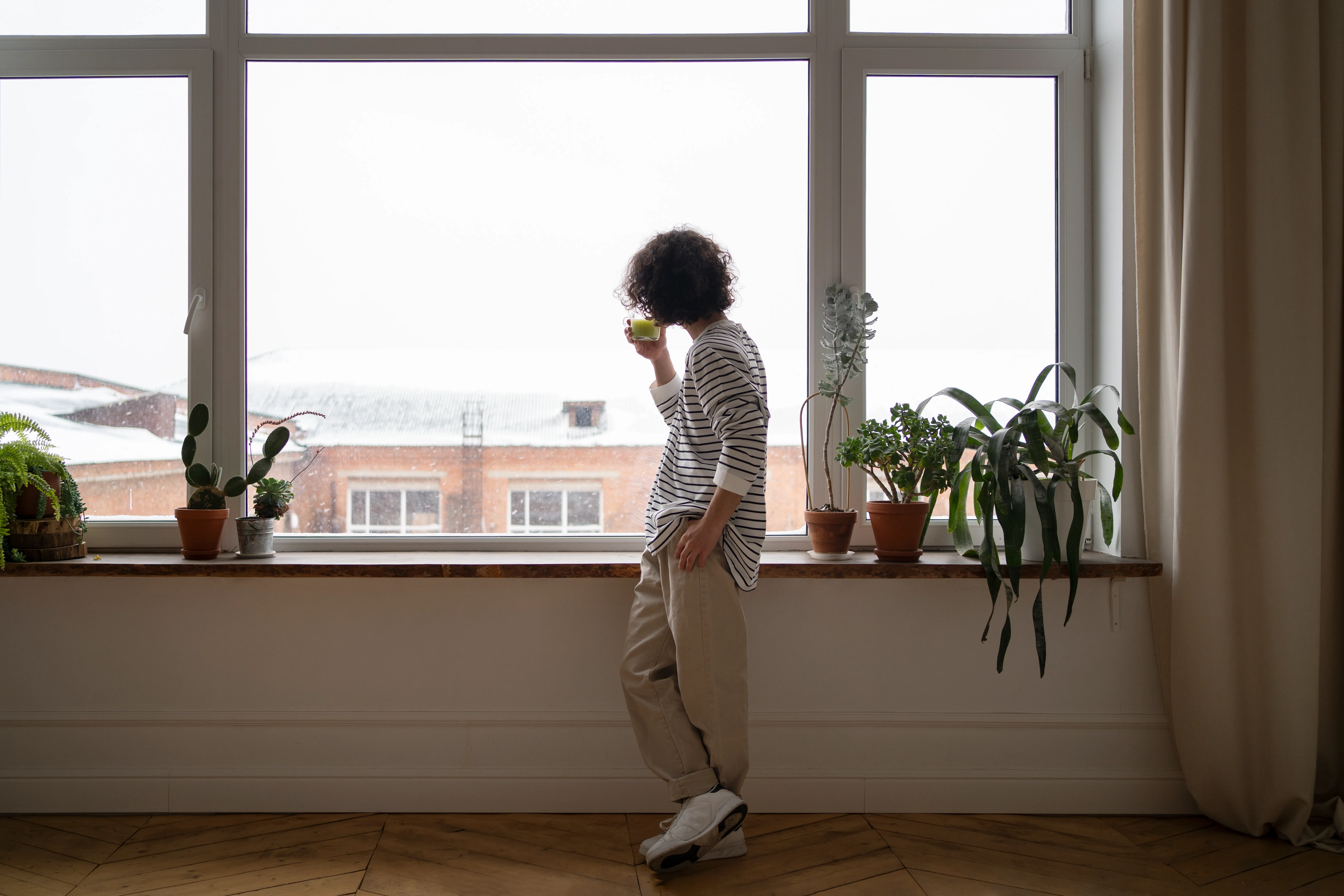Woman holding a warm drink beside a large window in a bright room during winter