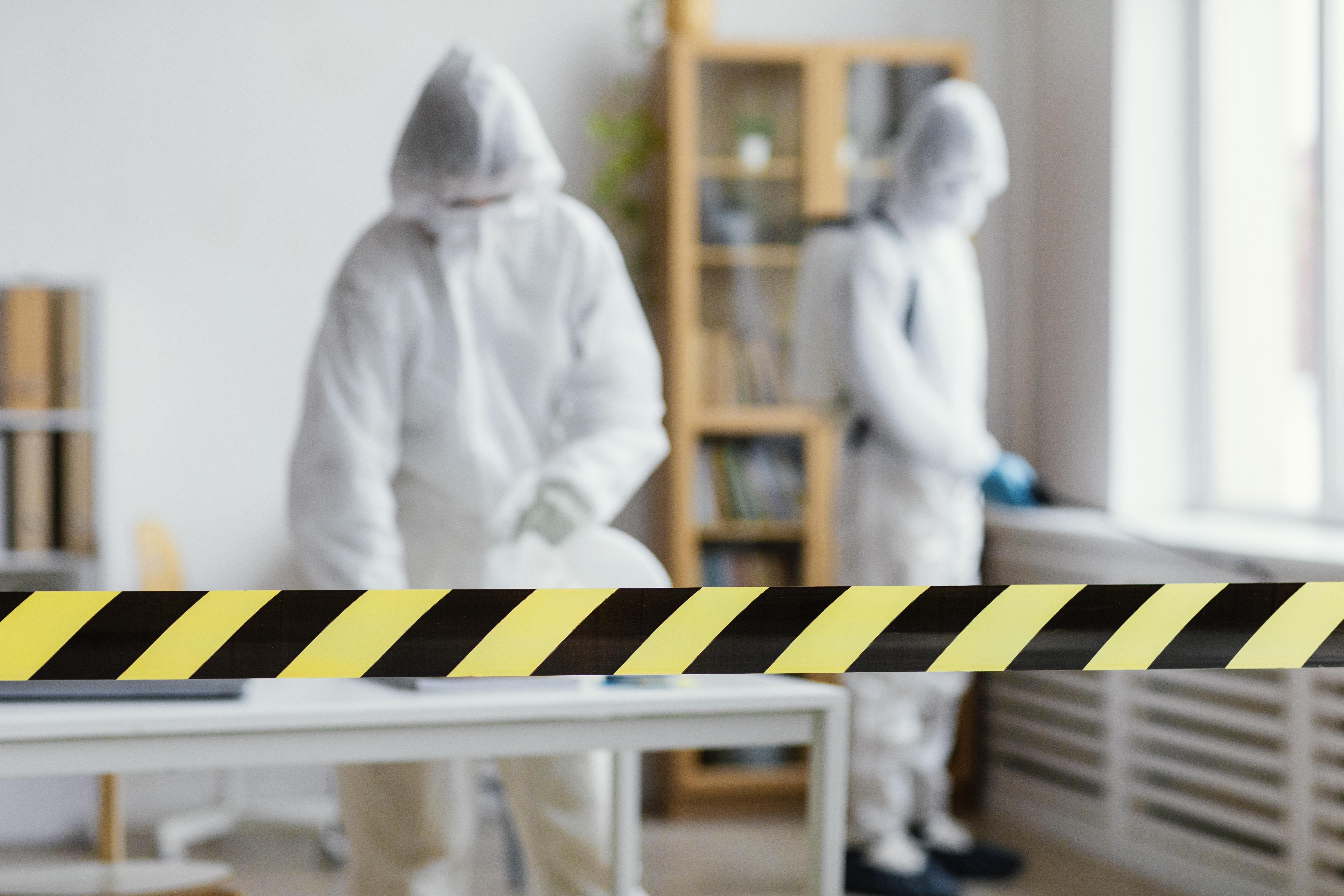 Two workers in protective suits and masks behind black and yellow safety tape indoors